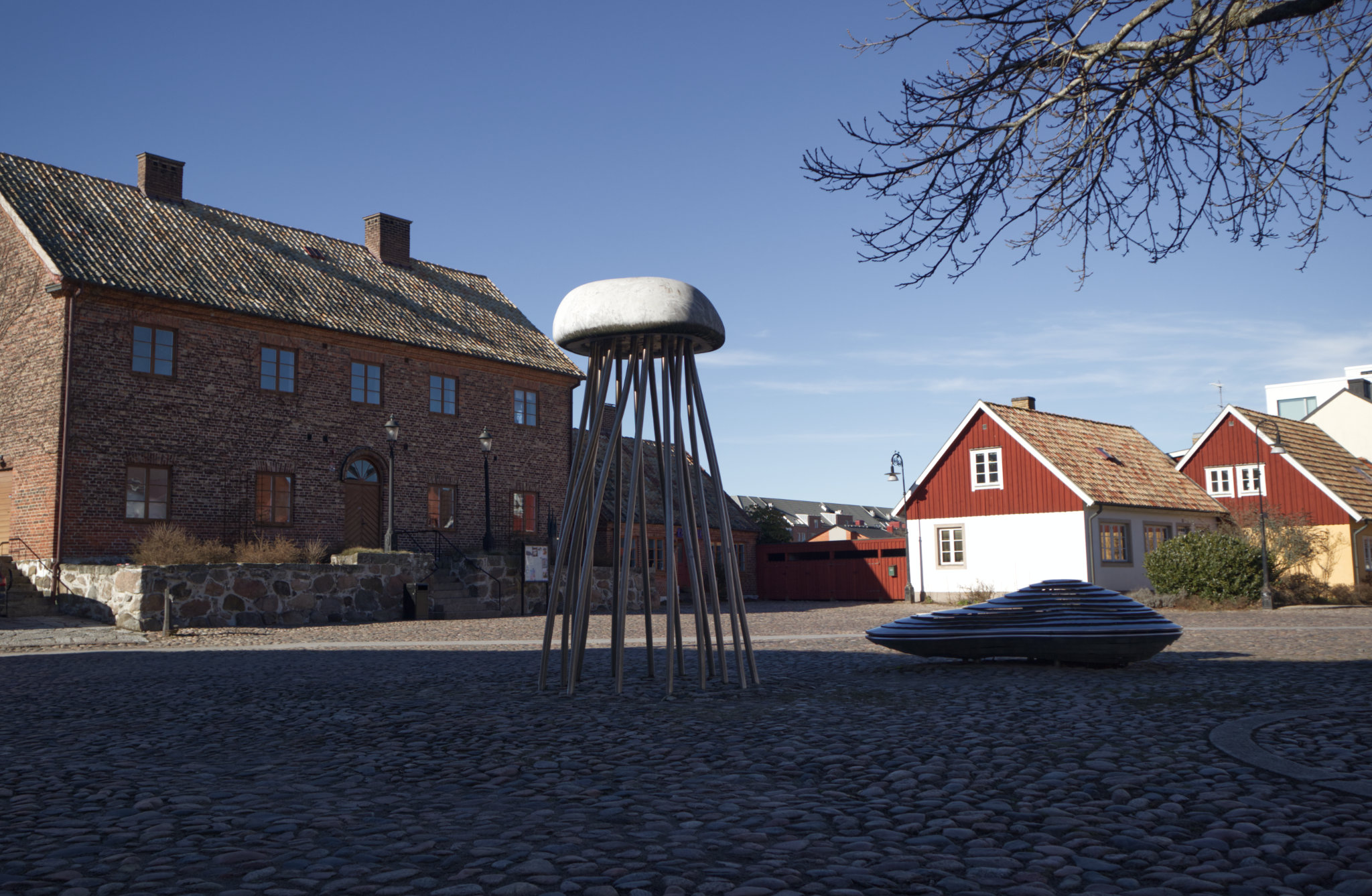 Bilden visar ett torg eller en öppen plats med kullersten i förgrunden. Till vänster i bilden står en stor tegelbyggnad med takpannor och flera fönster. Framför tegelbyggnaden finns en stenmur. I mitten av bilden finns en stor skulptur som liknar en