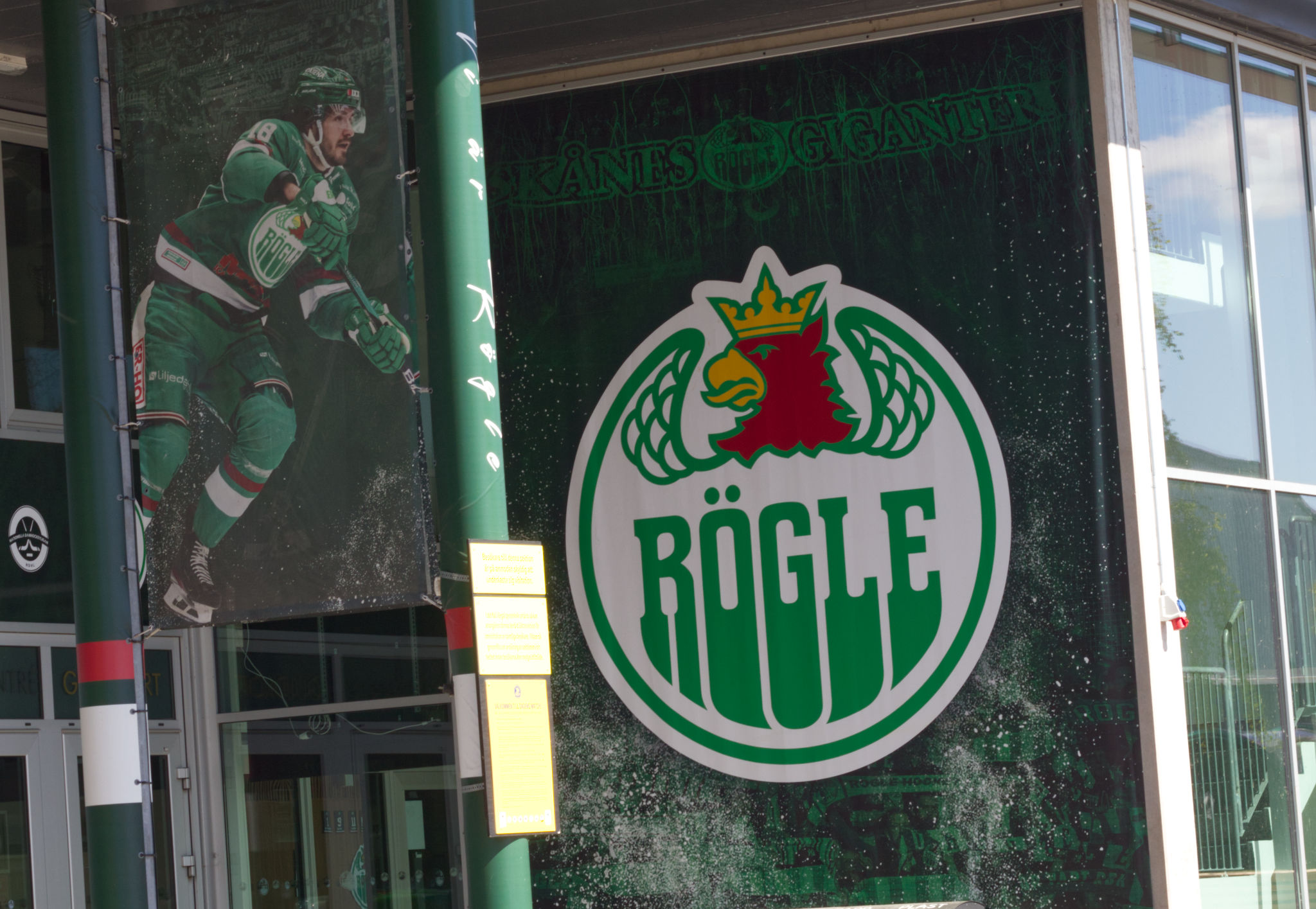 Bilden visar delar av en byggnadsfasad med två dörrar och en stor banderoll. Till vänster hänger en banderoll med bilden av en ishockeyspelare i färgen grön, medan den högra banderollen visar Rögle BK:s logotyp och texten "SKÅNES GIGANTER". Logotypen