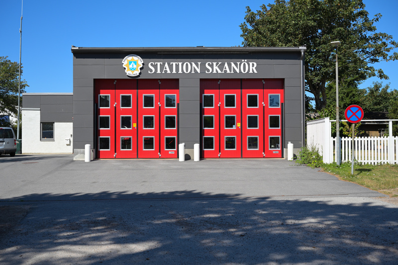 Bilden visar fasaden på en byggnad med texten "STATION SKANÖR" i stora vita bokstäver ovanför två stora, röda portar. Mellan texten och portarna finns en emblem som är svår att tyda. Byggnadsfasaden är grå med en vit sida till vänster. Framför byggna