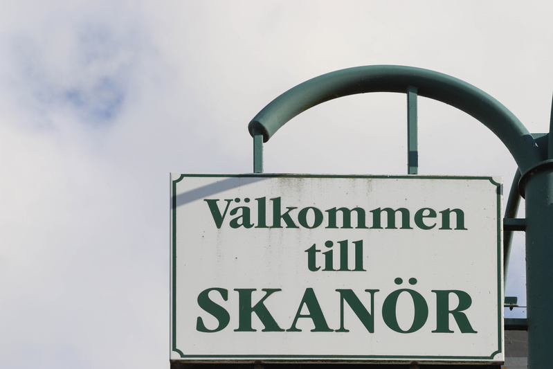Bilden visar en vit skylt med mörkgröna kanter och texten "Välkommen till SKANÖR" i mörkgröna versaler och gemener. Skylten är fäst vid en mörkgrön stolpe eller ram, varav en böjd del syns i bildens övre högra hörn. I bakgrunden syns en ljus himmel m