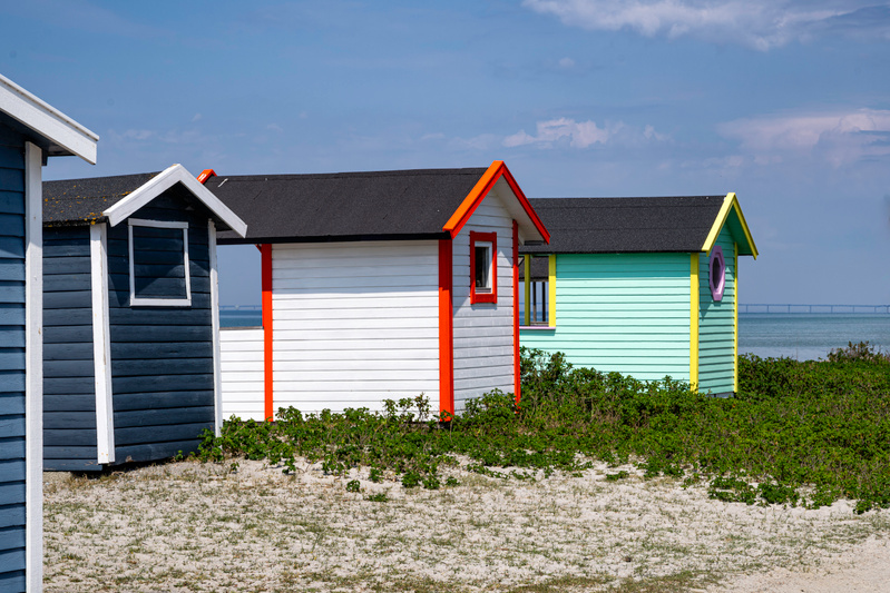 Bilden visar tre små färgglada badhytter eller bodar på en sandig strandbank. Till vänster syns en mörkblå hytt med vita detaljer och svarta tak. I mitten står en vit hytt med röda och orangea detaljer, inklusive fönsterramar och takkant. Till höger