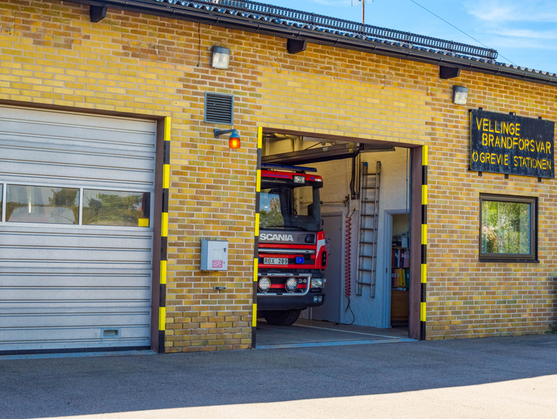 Bilden visar utsidan av en brandstation byggd i gult tegel under dagtid. Stationen har två garageportar med gula och svarta varningstejper längs kanterna, varav en är stängd och den andra är öppen. I den öppna porten syns fronten av en röd brandbil a