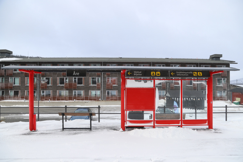 Bilden visar ett landskap täckt av snö. I förgrunden till vänster står ett rött busskurstak med en röd stolpe och en svart bänk delvis täckt av snö. Till höger syns en röd och vit busskur med text på svenska som indikerar 'Väntsal Biljetter' och 'Ers