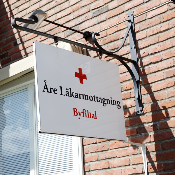 Bilden visar en vit skylt med texten "Åre Läkarmottagning Byfilial" skrivet i svart och röd färg. Ovanför texten finns en röd symbol som liknar ett kors. Skylten hänger från en mörk metallkonstruktion fäst på en tegelvägg. Till vänster om skylten syn