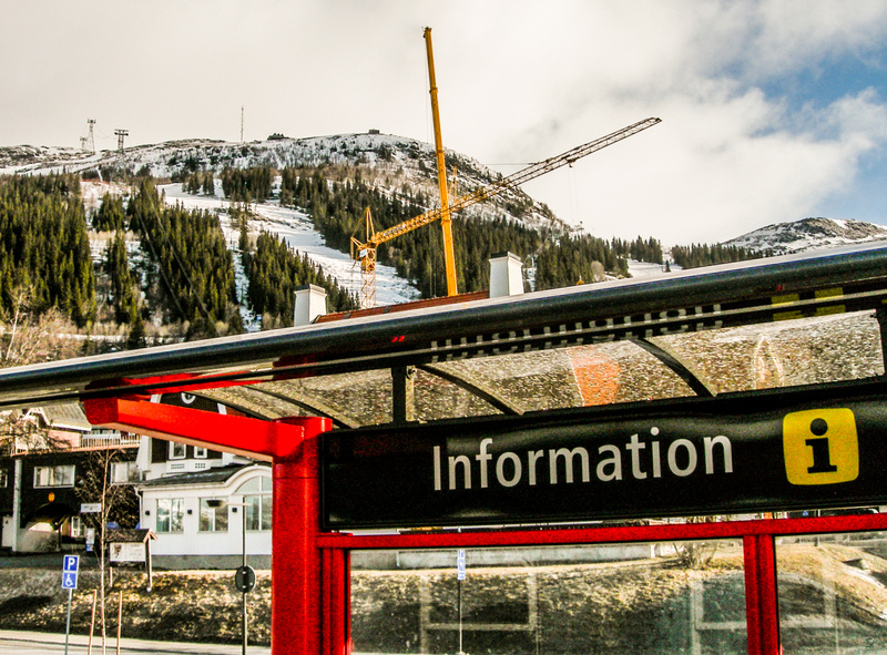 Denna bild visar en informationsskylt i förgrunden, som ingår i en röd och svart konstruktion med ett transparent tak. I bakgrunden syns ett snötäckt bergslandskap med täta barrskogar och delvis synliga skidbackar. Flera lyftkranar, varav en stor gul