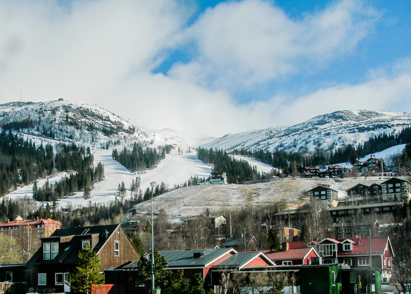 Bilden visar ett snötäckt bergslandskap med en skidbacke och skog. Längst ner i bilden syns en grupp hus med olika utseende, inklusive trähus med mörka tak och byggnader med röda tak. Vissa hus har stora fönster och balkonger. På bergssidan finns spå