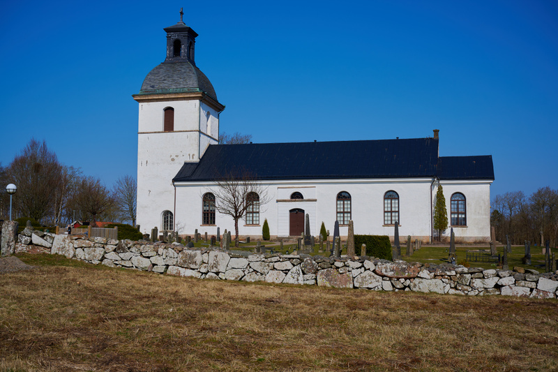 En vit kyrka med ett svart tak och en svart kupol på tornet står under en klarblå himmel. Kyrkan har en dörr och flera fönster med spröjs som kan vara spetsbågiga eller rundbågiga. Framför kyrkan syns en kyrkogård med flera gravstenar och en stenträd