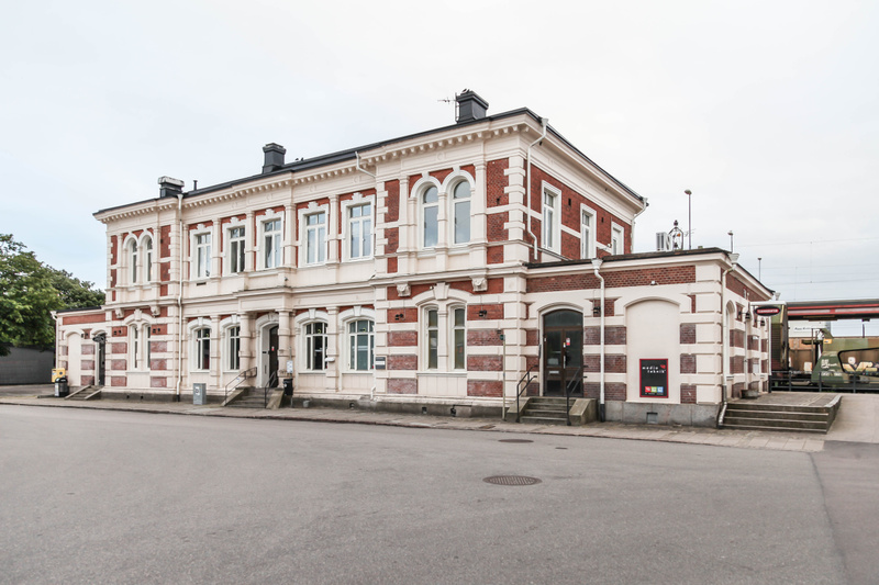 En stor, tegelröd byggnad med vita dekorationer står i centrum av bilden under en gråmulen himmel. Byggnaden är i två våningar på den vänstra och mellersta delen, medan den högra delen är lägre. Fasaderna har en kombination av exponerat tegel och vit