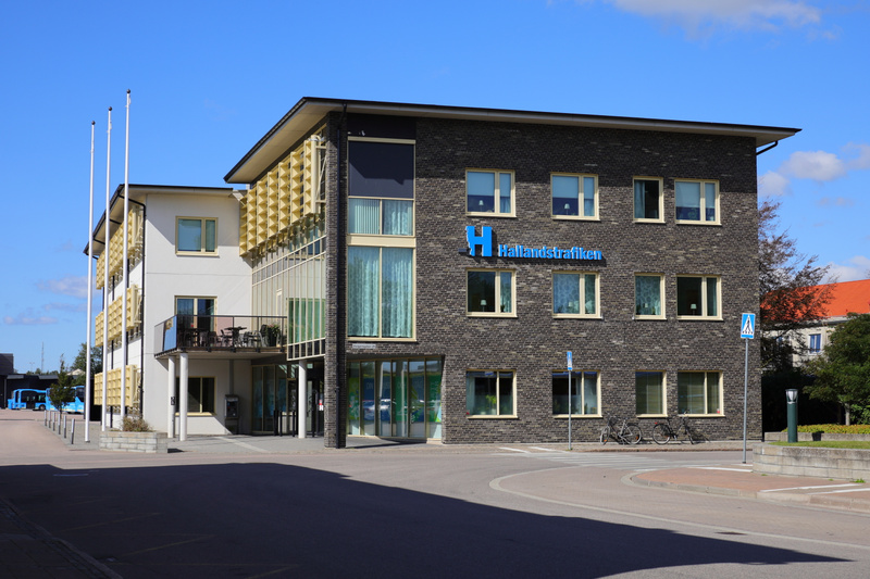 En modern byggnad med en fasad av mörk tegel och ljus puts. På den mörka tegelfasaden syns skylten 'Hallandstrafiken' i blå bokstäver. Byggnaden har flera fönster med ljusa ramar och vissa fönster på den ljusa putsfasaden har balkonger med svarta räc