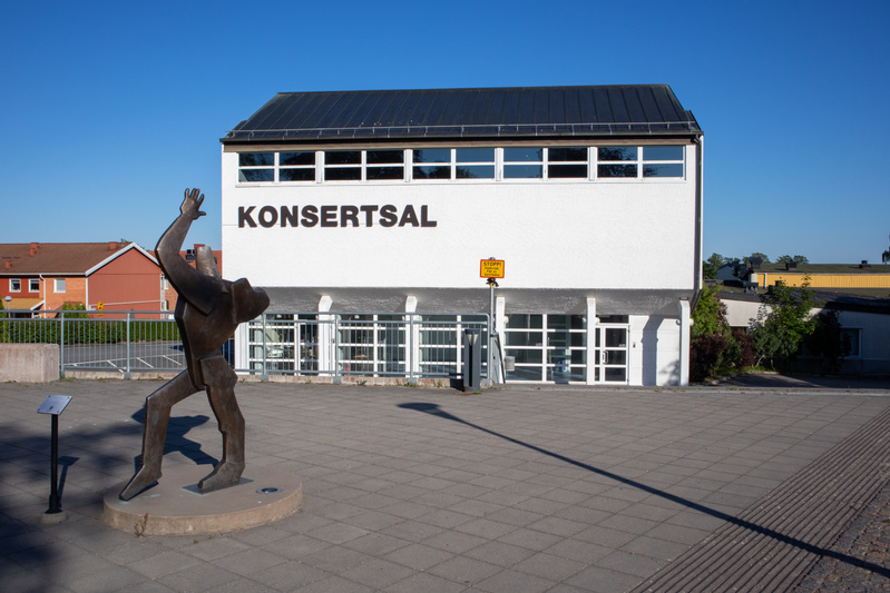 Bilden visar fasaden på en vit byggnad med texten "KONSERTSAL" i stora svarta bokstäver. Byggnaden har ett mörkt tak och ett långt fönsterband under takfoten, samt flera fönster längs den nedre delen. Framför byggnaden finns ett stenlagt torg med en