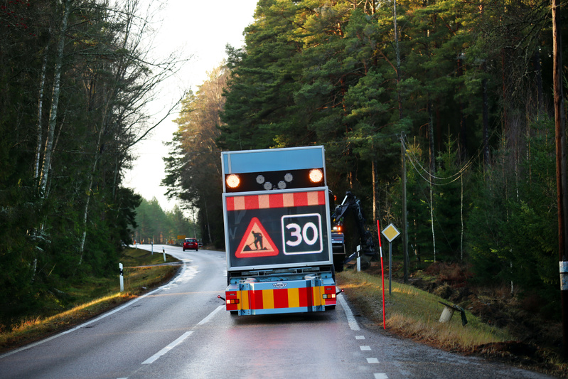En vägscen från baksidan av ett fordon med en stor digital skylt. På skylten syns ett varningsmärke för vägarbete (triangel med man som gräver) och en hastighetsbegränsning på '30'. Skylten har även blinkande orange lampor och en reflekterande röd- och vitrandig bakgrund. Vägen är en asfalterad landsväg som svänger svagt åt vänster framåt i bilden. Till vänster och höger om vägen växer träd, främst barrträd. Längre bort på vägen syns en röd bil. Dagsljus med solljus som lyser upp trädtopparna. Det finns vita streckade linjer som markerar vägkanten och en heldragen vit linje i mitten av vägen. En gul reflekterande skylt syns också bredvid vägen till höger.