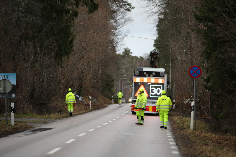 Bilden visar en väg med flera personer i gul varselklädsel. Två av personerna har ordet 'VAKT' tryckt på ryggen och står vid en lastbil med en digital skylt som visar hastighetsbegränsningen '30' och en varningssymbol. En tredje person i liknande klä