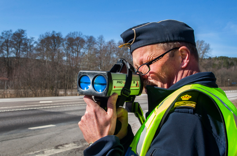 En person i mörkblå uniform och gul reflexväst håller och tittar igenom en handhållen hastighetsmätare. Personen har glasögon och en mörkblå keps med en guldig emblem. Hastighetsmätaren, märkt "PRO LASER III", har två blåaktiga linser riktade framåt.