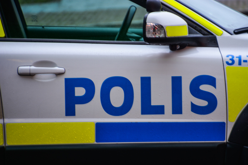Bilden visar närbilder på den högra sidan av en svensk polisbil. Huvudfokus ligger på mitten av dörren, där det blåa ordet "POLIS" syns tydligt mot den vita bakgrunden, med gult och blått reflekterande material under texten. Det finns vattendroppar p