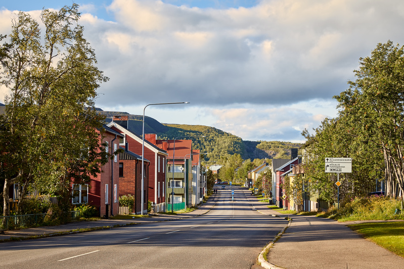 En gata i en stad med hus på båda sidor, under en delvis molnig himmel. Husen på vänster sida av gatan är mestadels röda och tegelröda, med några mindre byggnader och träd framför. På höger sida av gatan syns en blandning av hus i olika färger, samt