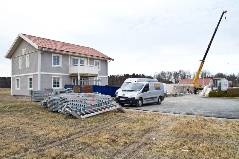 Bilden visar en byggarbetsplats utomhus under dagtid. I förgrunden syns ett gräsbevuxet fält med torrt gräs. Till vänster i bilden står ett nybyggt tvåvåningshus i grå fasad med vita fönsterkarmar och en röd takpanna. Huset har en balkong på andra vå