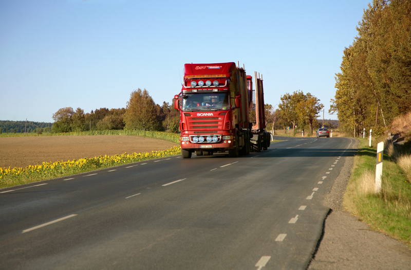 En röd timmerbil av märket Scania kör på en asfalterad landsväg under dagtid. Vägen har vita streckade linjer. Till vänster om vägen syns en jordbruksmark som är delvis täckt av solrosor, och längre bort skog. Till höger om vägen finns gräs och träd