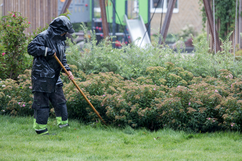 Bilden visar en person iklädd mörka regnkläder, inklusive en jacka med huva och byxor med reflekterande grön-gula detaljer på benen. Personen håller i ett långt träskaft, möjligtvis en kratta, och rengör gräsmattan från löv och växtrester. I bakgrund