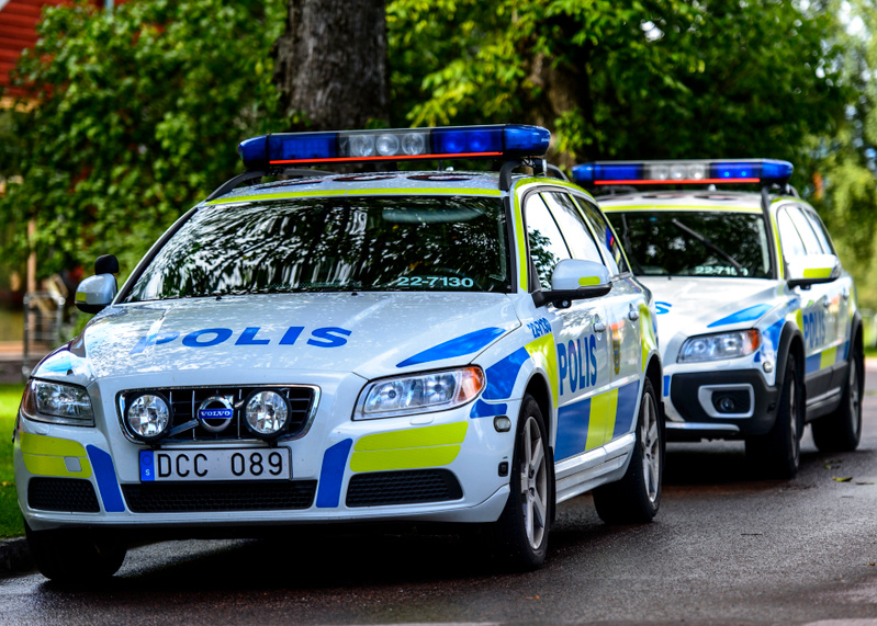 Bilden visar två vita svenska polisbilar, av modellen Volvo, parkerade på en asfalterad yta utomhus. Den främre polisbilen är tydligt synlig med texten "POLIS" i blått på huven och sidorna, samt blå och gula rutor. Takräcket är utrustat med blåa larm