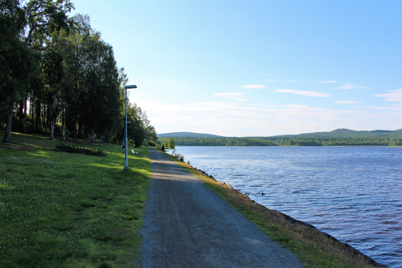 Bilden visar en grusväg längs ett vattendrag under dagtid. Till vänster om grusvägen finns en gräsmatta som övergår i en skog fylld med lövträd och barrträd. Längs grusvägen står flera lampstolpar och en vit bänk syns i bakgrunden. Vattendragets yter