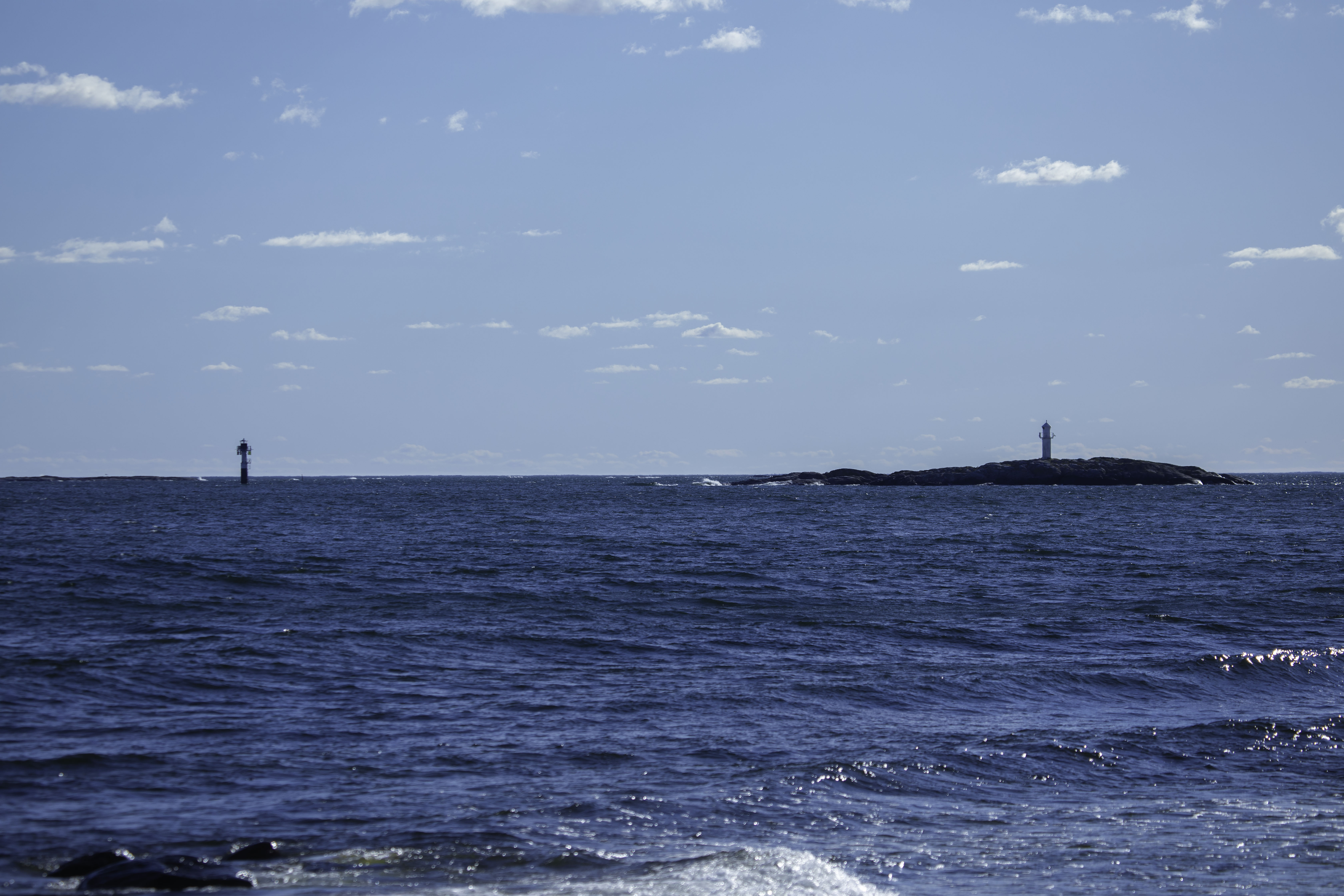 Bilden visar ett landskap med en klarblå himmel med spridda vita moln. I förgrunden syns mörkblått, vågigt vatten, med små vågor som skummar i nederkant. Mellan vattnet och horisonten syns två holmar eller skär med karga klippor. På den vänstra holme