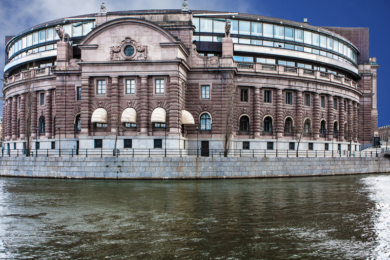 Bilden visar en stor byggnad i sten med en böjd form, sedd från andra sidan vattnet. Byggnadens nedre del är byggd av stora stenblock och dess fasad har detaljerade utsmyckningar, bland annat kolonner, fönster med markiser och ett centralt placerat e