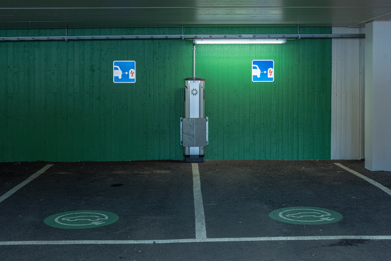 Bilden visar ett inomhusutrymme, sannolikt ett parkeringsgarage, med ett laddningsstation för elbilar. En grön trävägg sträcker sig över bildens centrum, och på den finns två blå skyltar med en vit symbol som indikerar laddning av elfordon. Mellan sk