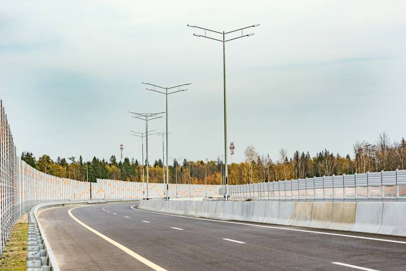 Bilden visar en asfalterad väg med två körfält som svänger åt vänster. Längs vägen finns långa grå bullerplank med orangea detaljer på båda sidor. På den högra sidan av vägen löper ett betongräcke närmast vägbanan, följt av bullerplank. På den vänstr