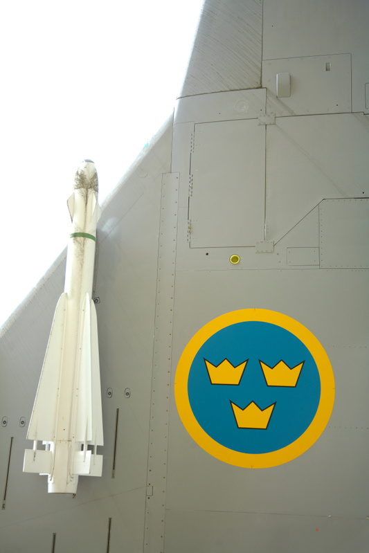 Bilden visar en detaljbild av ett militärflygplan. Till vänster i bilden syns en vit missil fastsatt på flygplanskroppen, med en grön rem runt sig. Flygplanets yta är grå med synliga nitar och panelfogar. På den grå ytan, nedre höger, är ett cirkulärt emblem målat. Emblemet består av en blå cirkel omgiven av en gul kant, och inuti den blå cirkeln finns tre gula kronor arrangerade med två upptill och en nedtill. Övre delen av bilden är överexponerad och lyser vitt.