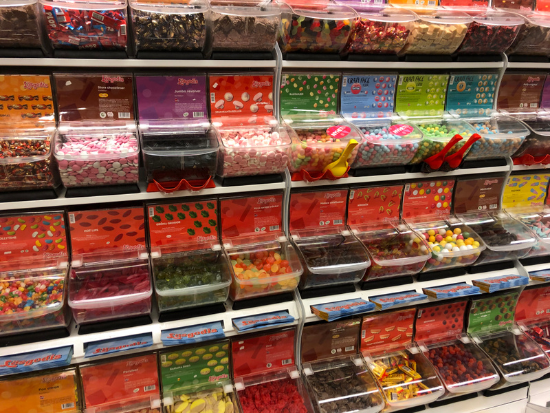 Bilden visar ett brett urval av lösgodis uppställt i genomskinliga plastbehållare på hyllor. Det finns flera rader med olika typer av godis, däribland chokladlinser i olika färger, gelégodis i varierande former och färger, samt godis med lakritssmak.