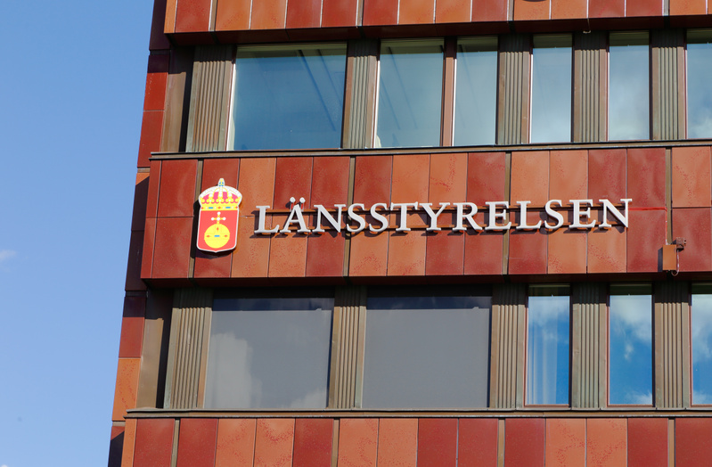 Bilden visar fasaden på en byggnad med texten "LÄNSSTYRELSEN" i vita versaler. Till vänster om texten finns en vapensköld i rött, gult och blått med en krona ovanför. Byggnadsfasaden är beklädd med bruna och rödbruna paneler i ett rutmönster. Det finns flera fönster i olika storlekar som reflekterar himlen. Byggnaden är fotograferad under dagtid med en klarblå himmel i bakgrunden. En del av byggnaden skyms av en skugga till vänster.