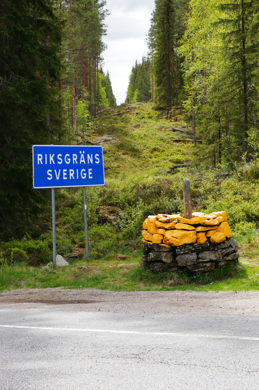 En bild som visar en svensk riksgräns i en skogsmiljö. I förgrunden finns en asfalterad väg med en vit linje. Till höger om vägen står en stenkonstruktion, delvis målad i en gulaktig färg, med en träpåle i mitten. Till vänster om vägen, på en gräsbevuxen yta, står en blå skylt med texten "RIKSGRÄNS SVERIGE" i vita versaler. Skylten är monterad på två silverfärgade stolpar. Området bakom skylten och stenkonstruktionen består av tät vegetation, inklusive gräs, buskar och många höga, barrträdsbetonade träd. Marken under vegetationen är ojämn och ser ut att ha inslag av stenar och mossbelagda ytor. Himlen i bakgrunden är ljus och molnig.