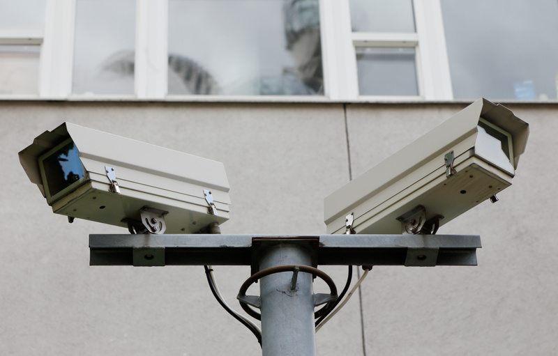 Bilden visar två övervakningskameror, monterade på en stolpe, med en grå byggnadsfasad och fönster med vita karmar i bakgrunden. Kamerorna är ljusgrå eller vita, rektangulära med en skyddande kåpa över själva linsen. De är vinklade åt varsitt håll och är fästa vid en horisontell bom som i sin tur sitter fast på en vertikal, metallfärgad stolpe. Vitlackerade fönsterkarmar och ett mönster som reflekteras i fönstren syns ovanför kamerorna och den grå väggen.