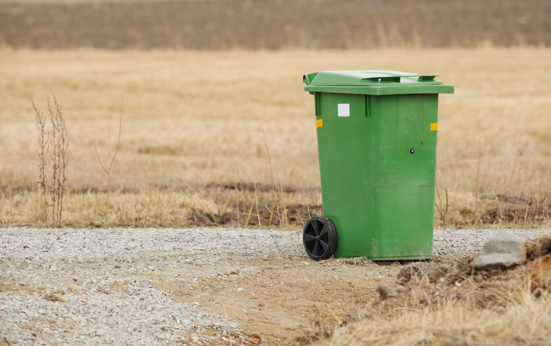 Bilden visar en grön soptunna på hjul placerad intill en grusväg eller grusgång. Tunnan har vita och gula klistermärken på sidan. Bakgrunden består av ett fält med torr, gulbrun vegetation och en horisontlinje som indikerar att det är ett öppet landskap. Marken närmast soptunnan är grusig och jordig med viss torkad vegetation. En mindre sten syns i bildens högra förgrund och viss torkad växtlighet i vänstra förgrunden.