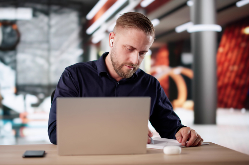En man med skägg sitter vid ett träbord och arbetar. Han har på sig en mörkblå skjorta och vita trådlösa hörlurar i öronen. Framför honom på bordet står en silverfärgad laptop som är öppen, men skärmen syns inte i bild. Till vänster om laptop finns e