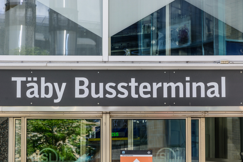 Bilden visar en skylt med texten "Täby Bussterminal" i vit text på en mörkgrå bakgrund. Skylten är fäst ovanför en glasfasad, delvis reflekterande grönskande träd och byggnaders inre. Ovanför skylten finns en fönsterrad med vinklade glaspartier. Neda