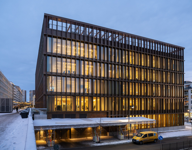 En stor, mörk byggnad med många upplysta fönster dominerar bilden. Byggnaden har en modern arkitektur med vertikala lameller som täcker fasaden. Framför byggnadens nedre del syns ett takliknande skydd med byggnadsställningar under, och bredvid detta