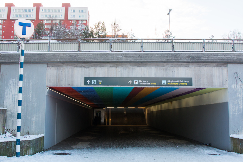 Bilden visar ingången till en tunnelbanestation eller gångtunnel i vintertid. Mark och ytor är täckta av snö. Ett betongöverhäng bildar ingången till en mörkare tunnel. Ovanför tunnelöppningen sitter en skylt med pilar och text på svenska och engelsk