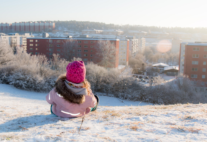 En person sedd bakifrån sitter på en snötäckt backe med en vid utsikt över ett bostadsområde. Personen bär en rosa mössa med mönster, en ljusrosa vinterjacka med pälskrage och reflekterande detaljer på ryggen. Jackan har också reflekterande detaljer