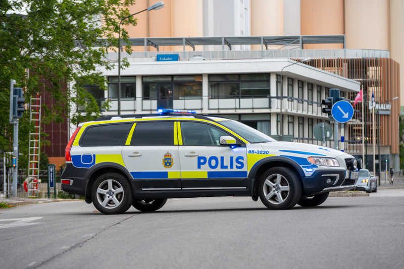 En svensk polisbil, en vit Volvo XC70 med blå och gula markeringar samt texten "POLIS" på sidan, står parkerad på en väg. Bilens blåljus är tända. I bakgrunden syns flera byggnader. En byggnad har stora fönster och en skylt som säger "Lokaler", medan