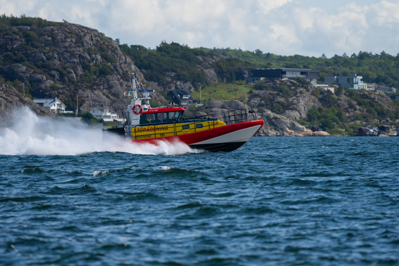 Bilden visar en röd, gul och vit sjöräddningsbåt som färdas hastigt över vatten. Båten skapar ett vitt stänk av vatten på sin vänstra sida. På båtens sida står texten "SJÖRÄDDNING" och nedanför "JOSEPHINE" i svarta bokstäver. En svensk flagga syns på
