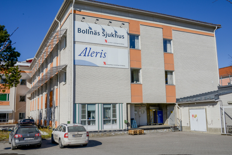 Bilden visar utsidan av en byggnad som identifieras som "Bollnäs Sjukhus" och "Aleris" genom stora skyltar på fasaden. Byggnaden är flervånings och har en kombination av ljus tegelstensfasad och orangefärgade paneler runt fönstren. Flera fönster kan