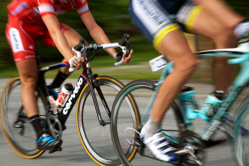 Bilden visar två cyklister i rörelse från sidan, tagna med en panoreringsteknik som ger en känsla av fart genom att bakgrunden är suddig medan cyklisterna är relativt skarpa. Den cyklist som är närmast kameran bär en röd cykeltröja och cykelshorts, o