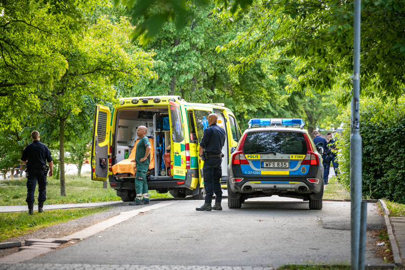 Bilden visar en stillbild i en utomhusmiljö, troligen på en gata eller parkeringsplats, omgiven av träd och grönska. I förgrunden syns en gul ambulans med öppen bakdörr, samt en mörkblå polisbil av kombi-modell med registreringsskylten 'WFS 835'. Blå