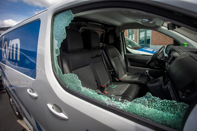 Bilden visar utsidan av en vit skåpbil med texten "KVM" och "ENTREPRENAD AB" i blått på sidan. Fönstret på passagerarsidan är krossat, och glasbitar ligger kvar i fönsterkarmen samt på de svarta sätena inuti bilen. Inredningen i fordonet, inklusive r
