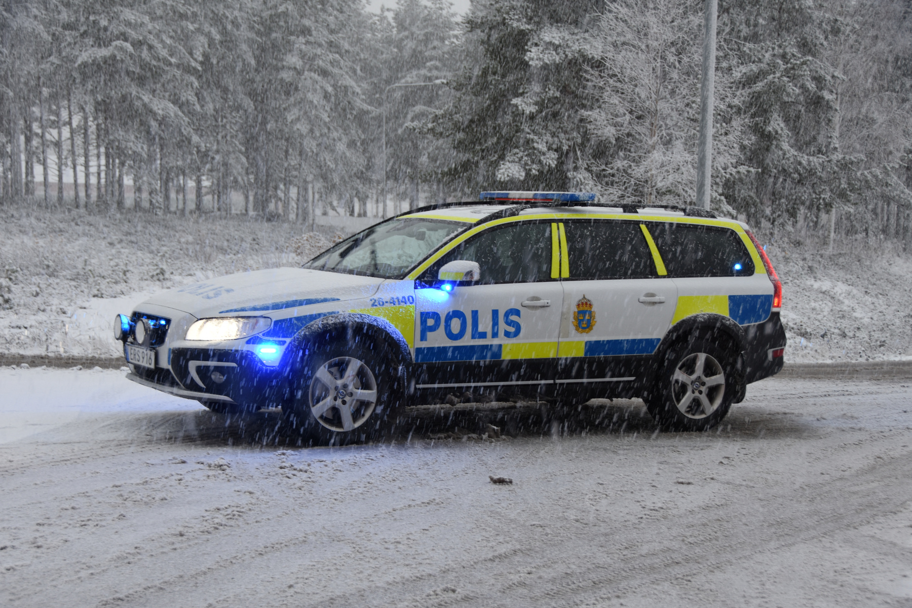 En svensk polisbil av modellen Volvo V70 står på en snötäckt väg i lätt snöväder. Bilen är huvudsakligen vit med blå och gula rutor och texten "POLIS" i blått på sidan. På taket syns blå utryckningsljus, och framtill är bilens strålkastare tända till