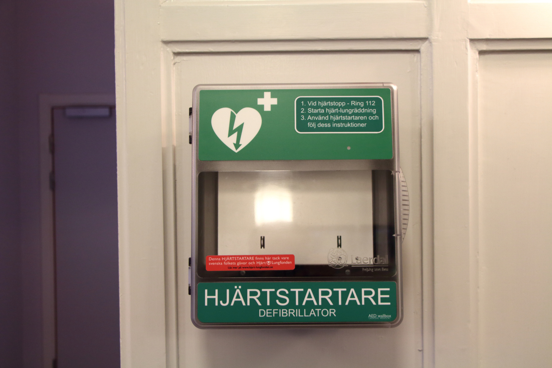 Bilden visar en hjärtstartare, en defibrillator, monterad på en vit dörr eller vägg. Hjärtstartaren är innesluten i ett vit- och grönfärgat skåp med en transparent front. På skåpets ovansida finns en grön skylt med en vit symbol föreställande ett hjä
