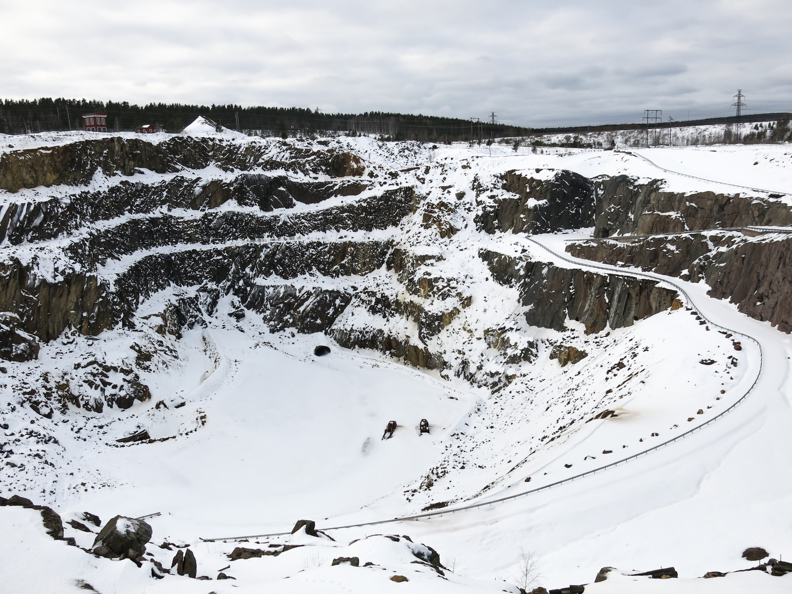 En vidvinkelbild som visar en stor, snötäckt dagbrottsgrop under en molnig himmel. Gropen har flera terrasserade klippväggar som är täckta av snö. En slingrande väg med skyddräcke kan ses längs gropens sida. I botten av gropen, som också är täckt av