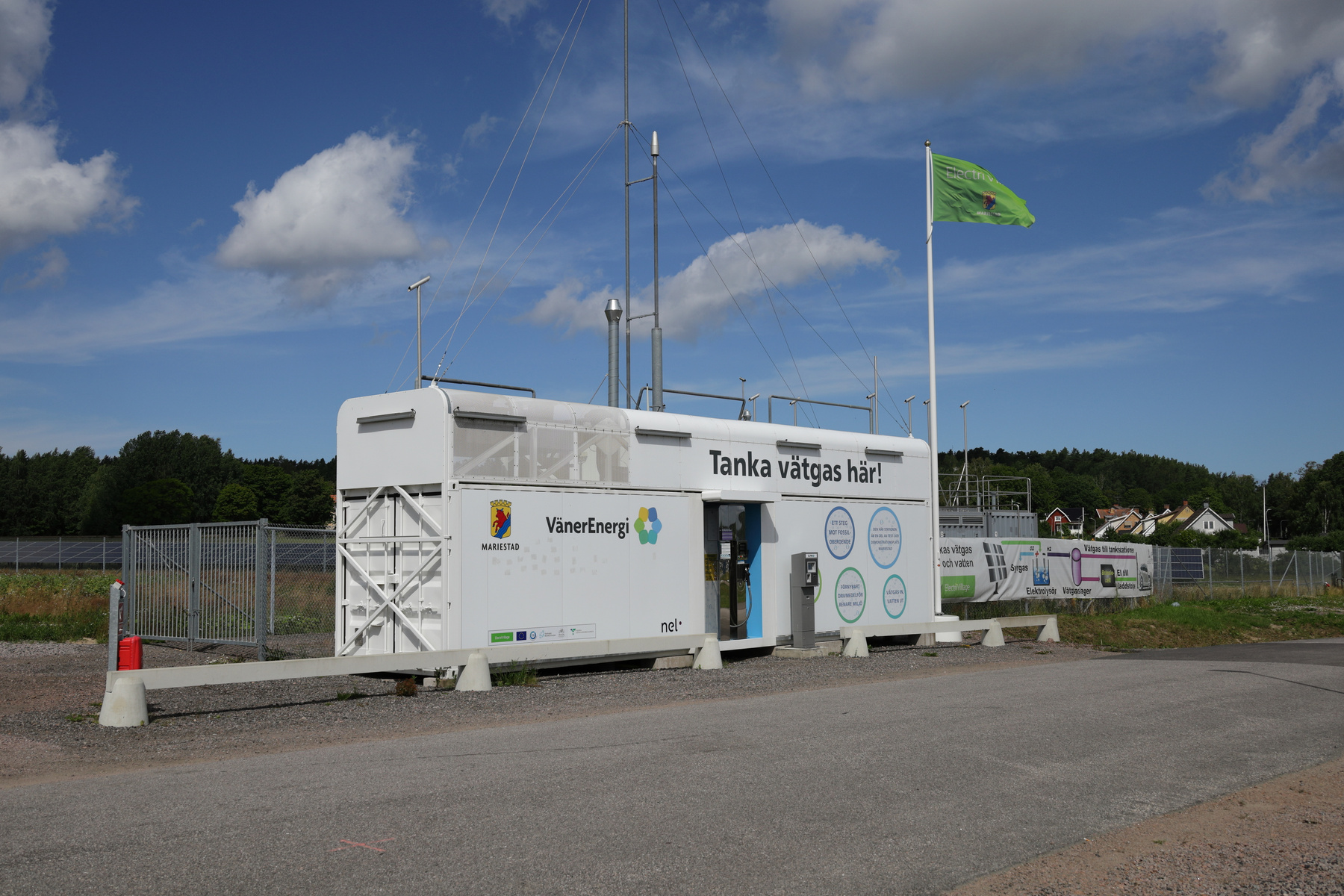 En modern vätgasstation står placerad utomhus under en klarblå himmel med spridda moln. Stationen består av en stor vit containerliknande byggnad med texten "Tanka vätgas här!" på sidan. Flera skyltar och logotyper såsom "VänerEnergi" och "Mariestad"