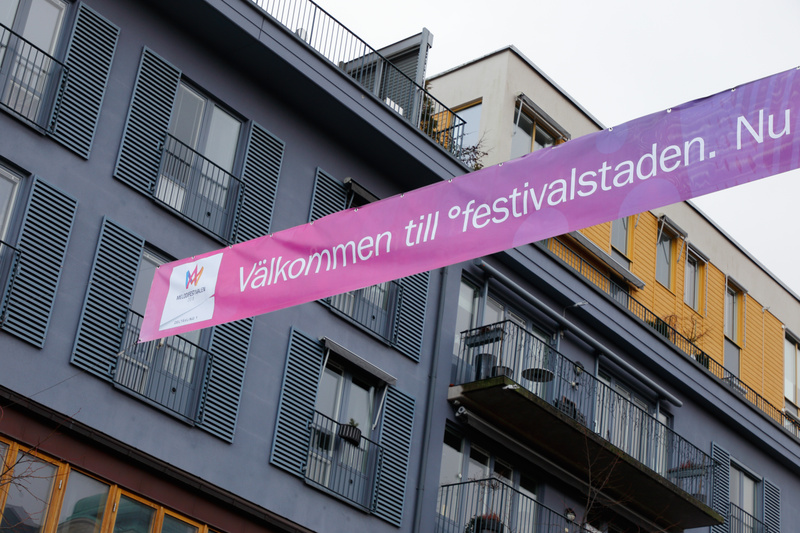 Bilden visar en ljuslila banderoll som sträcker sig horisontellt över bildytan, fäst mellan två byggnader. På banderollen står texten "Välkommen till °festivalstaden." med vita bokstäver. Till vänster på banderollen finns en logotyp med texten "MELLOFESTIVALEN" och en symbol. I bakgrunden syns delar av bostadshus med flera fönster och balkonger. Fönstren har grå luckor eller persienner. En byggnad är mörkgrå, medan en annan är gul, och en tredje har ljusa fasader. Molnigt väder syns i bakgrunden.